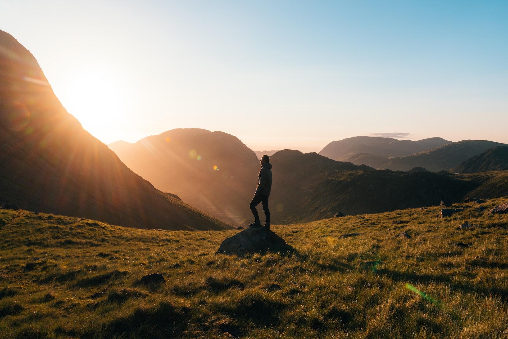 Man staying motivated by taking a deep breath of fresh air while standing on a rock on some grass in front of mountains during the sunset